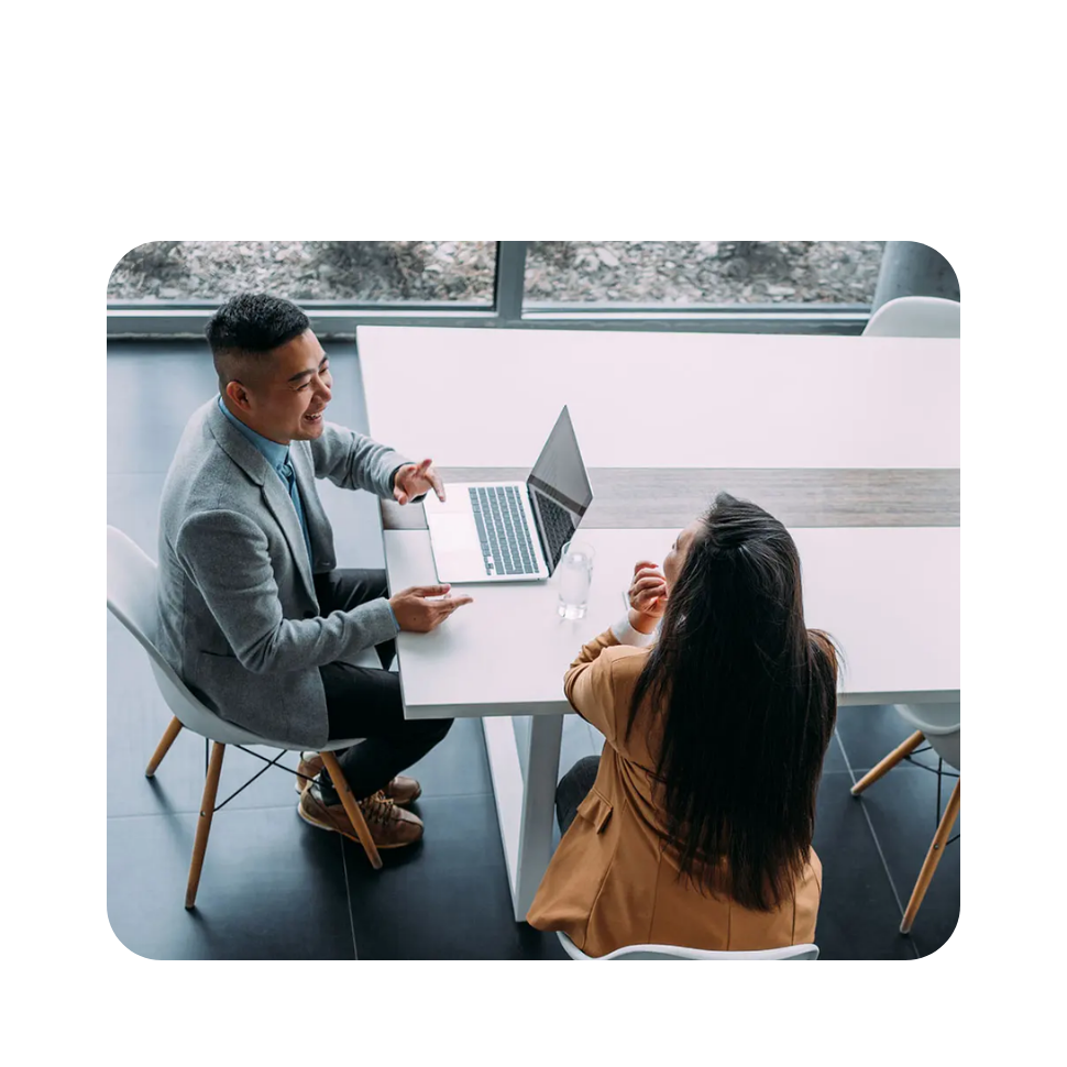 Two professionals having a discussion at a table with a laptop in a modern office setting with large windows.